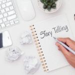 Man's hand writing word story telling on notebook, high angle view of screenwriter's desk for film, drama, musical and documentary.