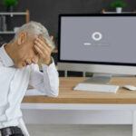 Unhappy, sad senior man sitting at office desk with computer with gear icon and progress bar on monitor, holding head in despair, waiting for software update to install or new operating system to load