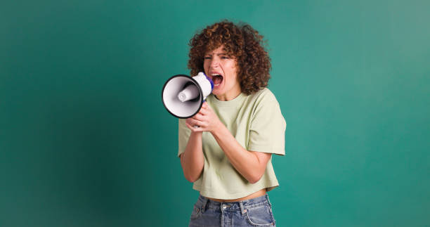 Cheerful young woman shouting on megaphone while standing against green background