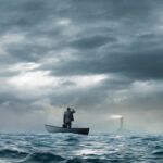 A beacon from a lighthouse beckons a stranded businessman as he stands in a small boat that floats under an ominous sky and choppy waters.