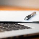 Laptop Computer on a Modern Wooden Business Desk with a Notepad and Pen in Unfocused Background.