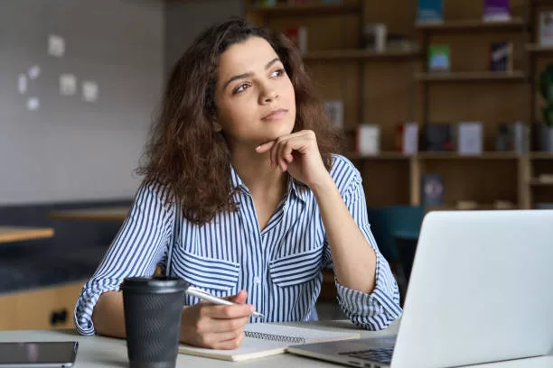 How To Work Through Writer's Block. - Young indian dreamy thoughtful student looking away working on laptop in office coworking space classroom. Hispanic student using computer for remote learning online training.